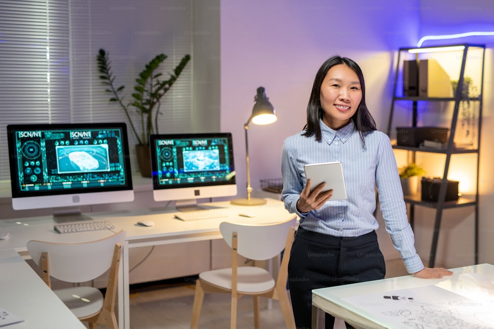 Porträt eines lächelnden attraktiven asiatischen Autodesigners in gestreiftem Hemd, der ein digitales Tablet in einem modernen Büro mit Computern hält