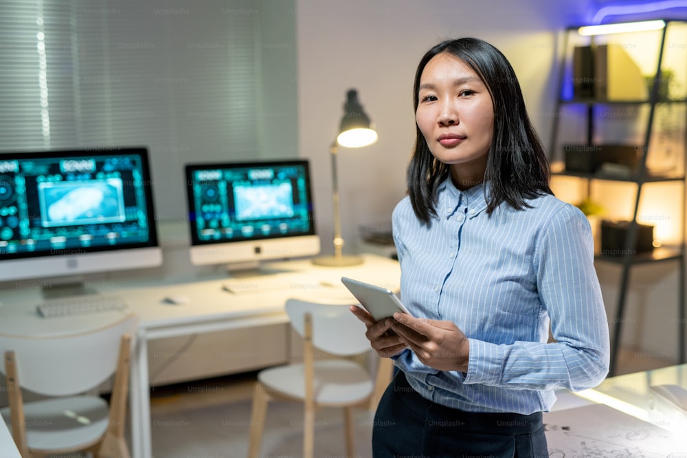 Retrato do conteúdo confiante jovem mulher asiática na camisa segurando tablet no escritório contemporâneo, ela trabalhando até tarde