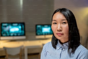Portrait of content confident young Asian businesswoman standing against computers in dark office