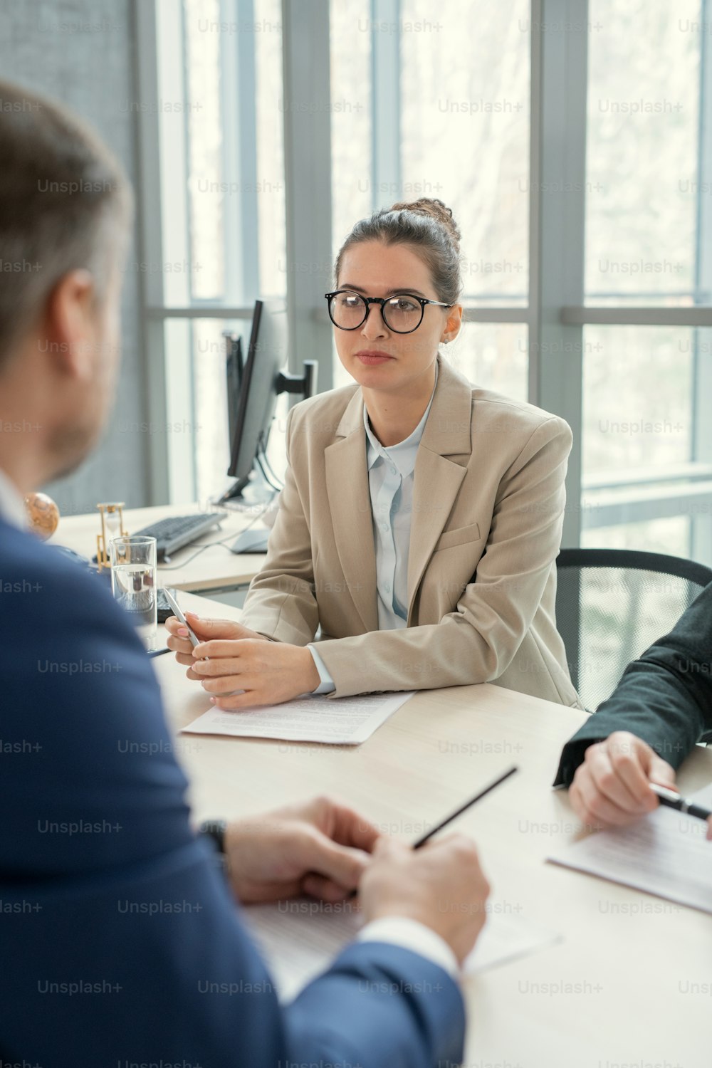 Content confident lady in eyeglasses sitting at table and holding pen while listening to colleagues at meeting
