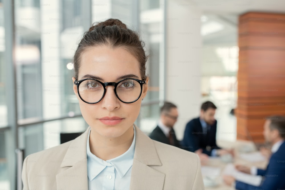 Portrait of confident attractive female attorney in round eyeglasses standing against colleagues discussing papers in background