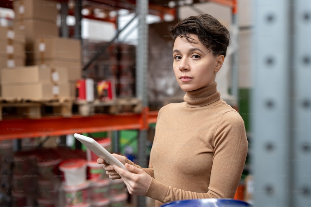 Portrait of serious supply manager with brown hair standing against warehouse frame and working with data on tablet