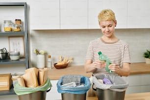 A woman holding plastic bottle over trash bin while sorting waste in the kitchen