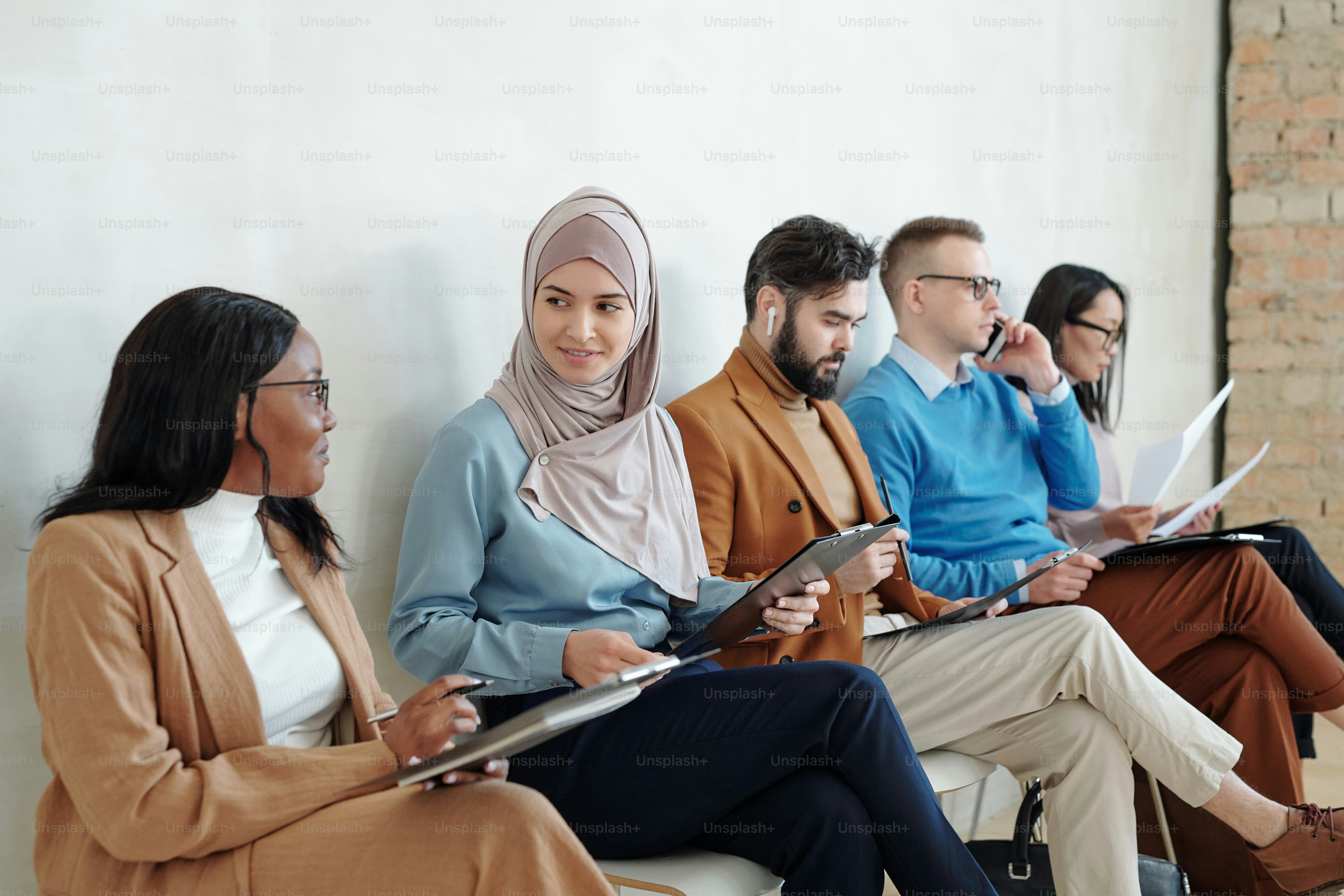 Smiling young Black and Arabian women sitting on chairs in corridor with other job candidates and discussing vacancy while being in line