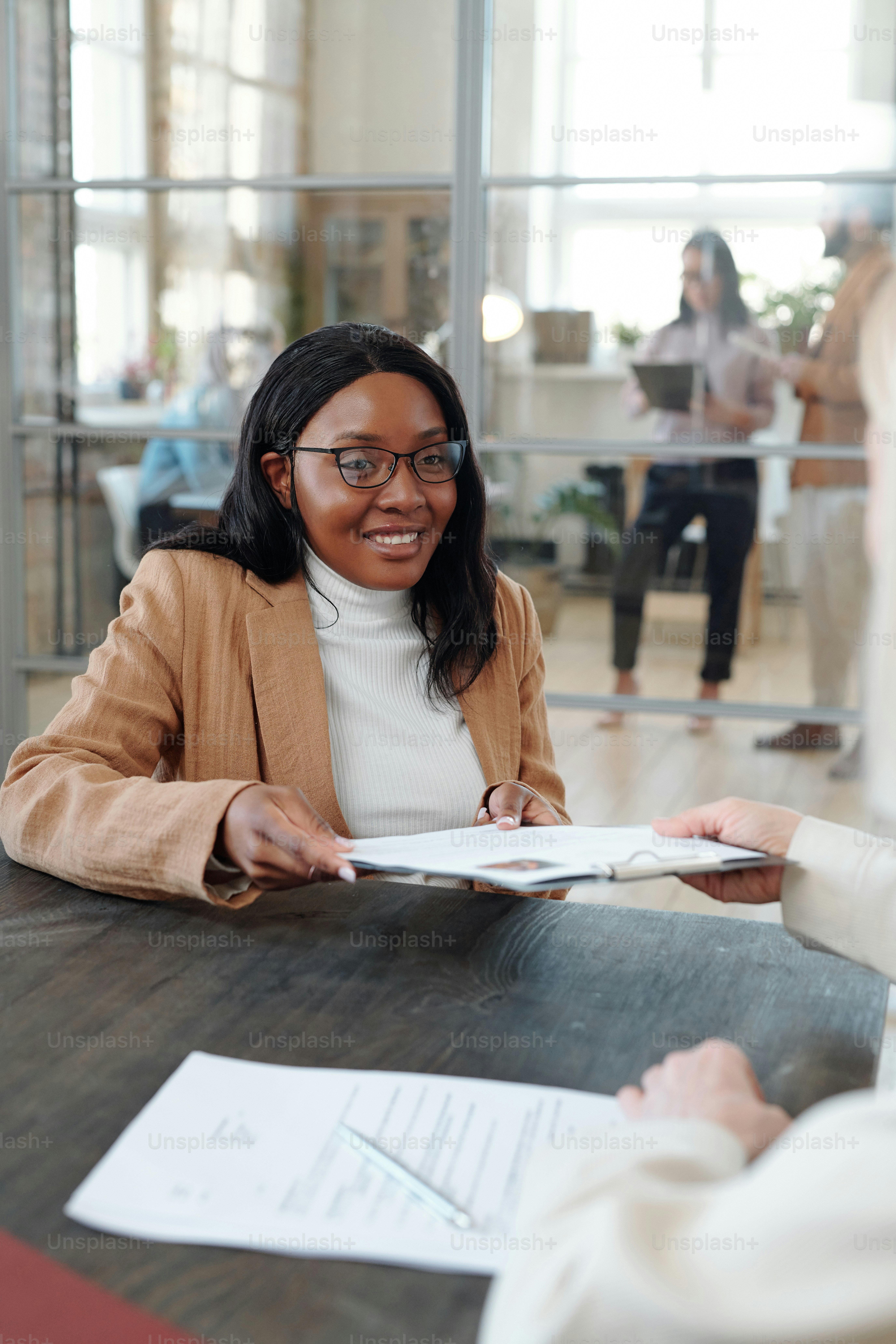Smiling young Afro-American woman in glasses sitting at table and giving her CV to HR manager during job interview
