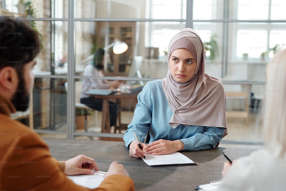 Giovane donna araba seria in hijab seduta al tavolo di fronte ai responsabili delle risorse umane e compilazione del questionario al colloquio di lavoro