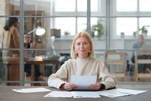 Portrait of content mature HR manager with blond hair sitting at table and reading CVs of candidates in modern open space office