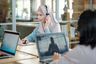 Serious young Arabian call center agent adjusting headset microphone while responding to customer using laptop in office