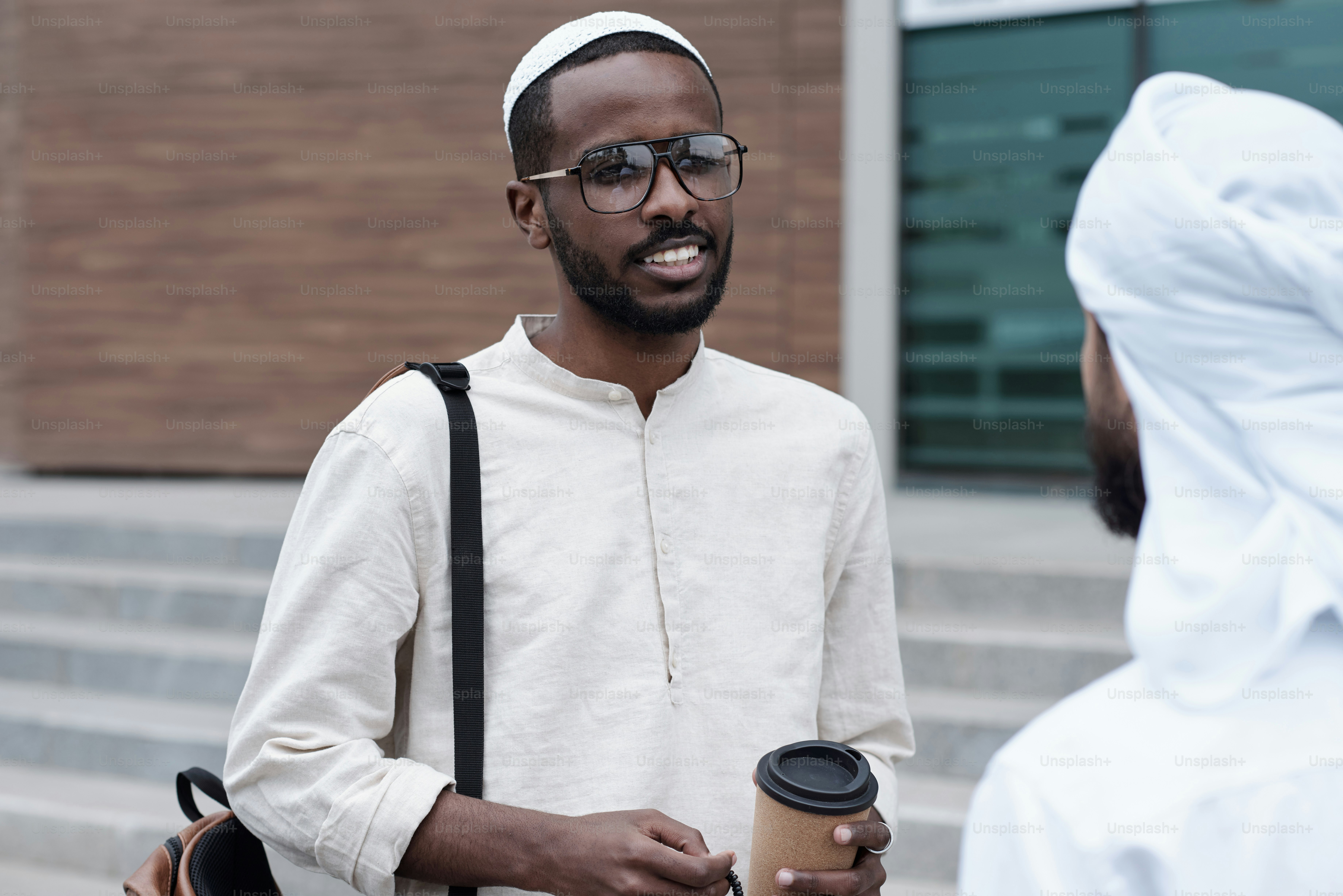Smiling young Black man with beard drinking coffee against modern office building and talking to Arabian colleague