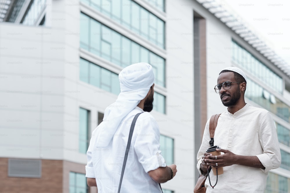 Content young multi-ethnic men in islamic clothes standing against modern building and talking about business outdoors