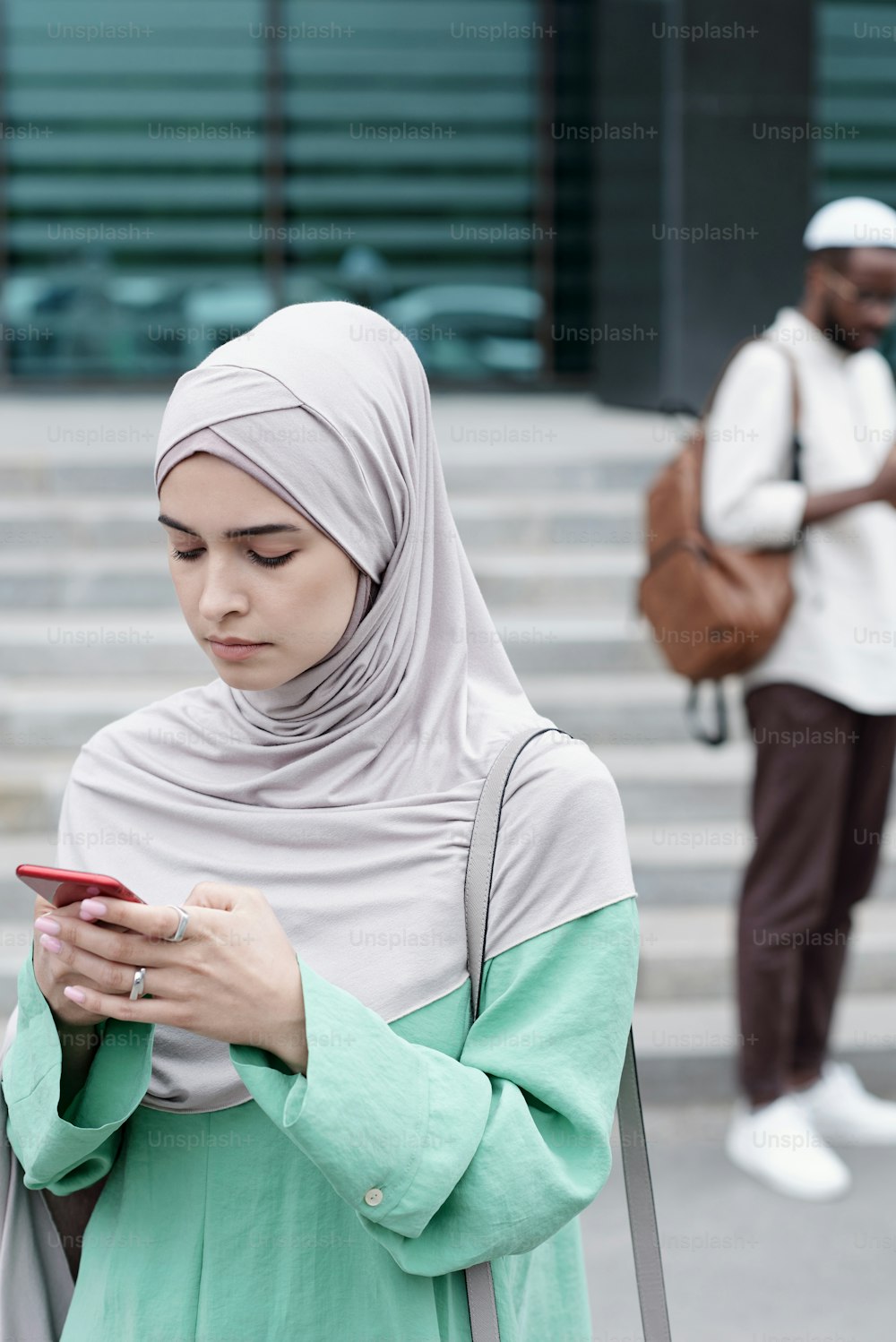 Ernsthafte junge muslimische Geschäftsfrau in Hijab und Nationaltracht benutzt Smartphone, während sie nach der Arbeit ein Taxi bestellt