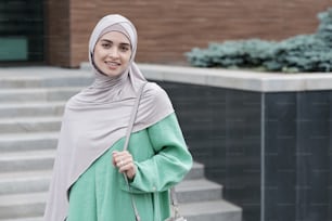 Portrait of positive modern young Islamic businesswoman holding handle of shoulder bag against contemporary office building