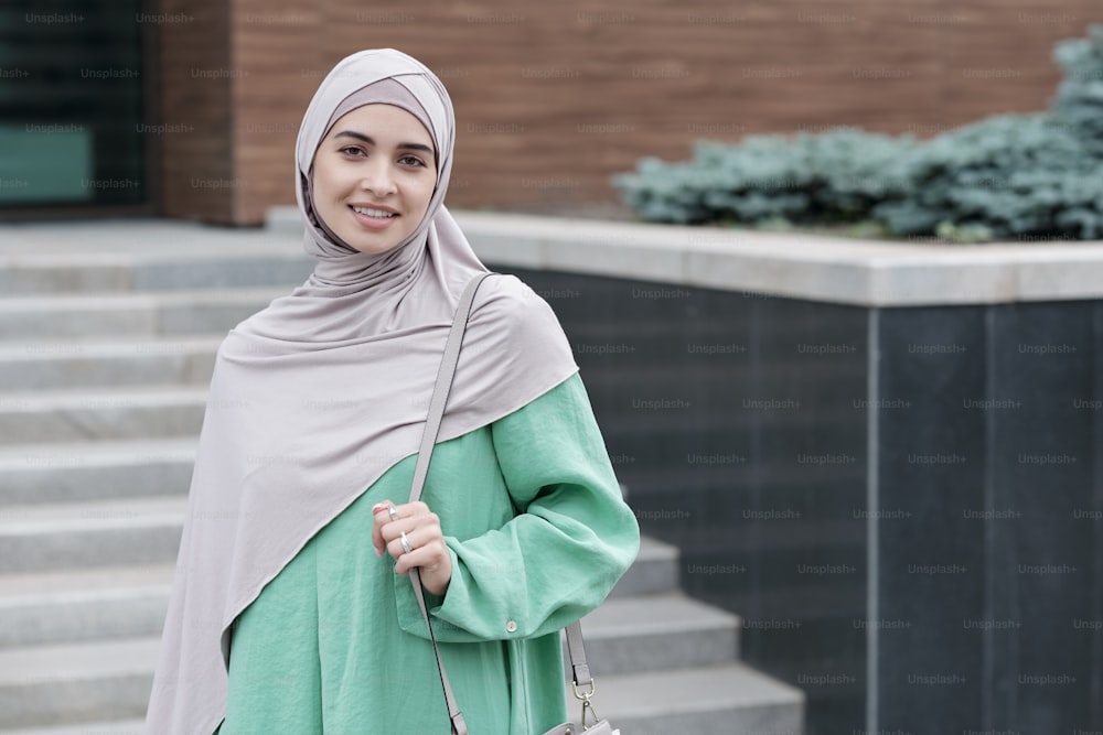 Portrait of positive modern young Islamic businesswoman holding handle of shoulder bag against contemporary office building