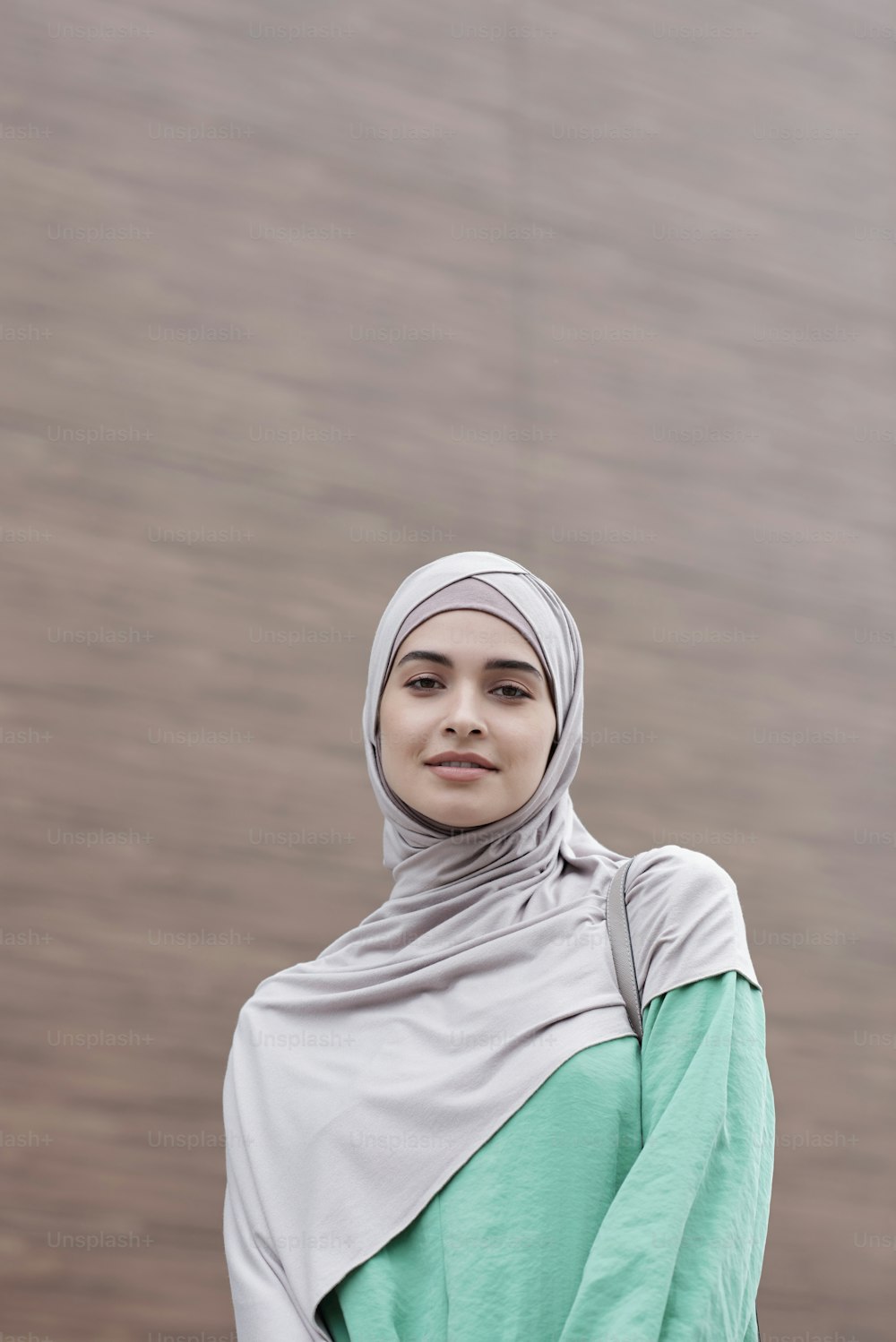 Porträt einer lächelnden schönen jungen muslimischen Frau in Hijab und grünem Kleid, die vor einem zeitgenössischen Gebäude steht