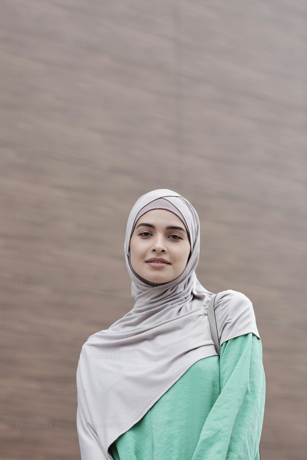 Portrait of smiling beautiful young Muslim woman in hijab and green dress standing against contemporary building