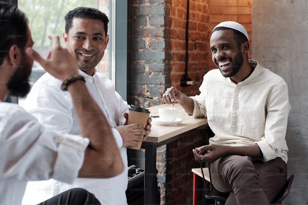 Groupe de jeunes amis musulmans joyeux buvant du café et riant à la blague de l’homme tout en se rassemblant dans un café