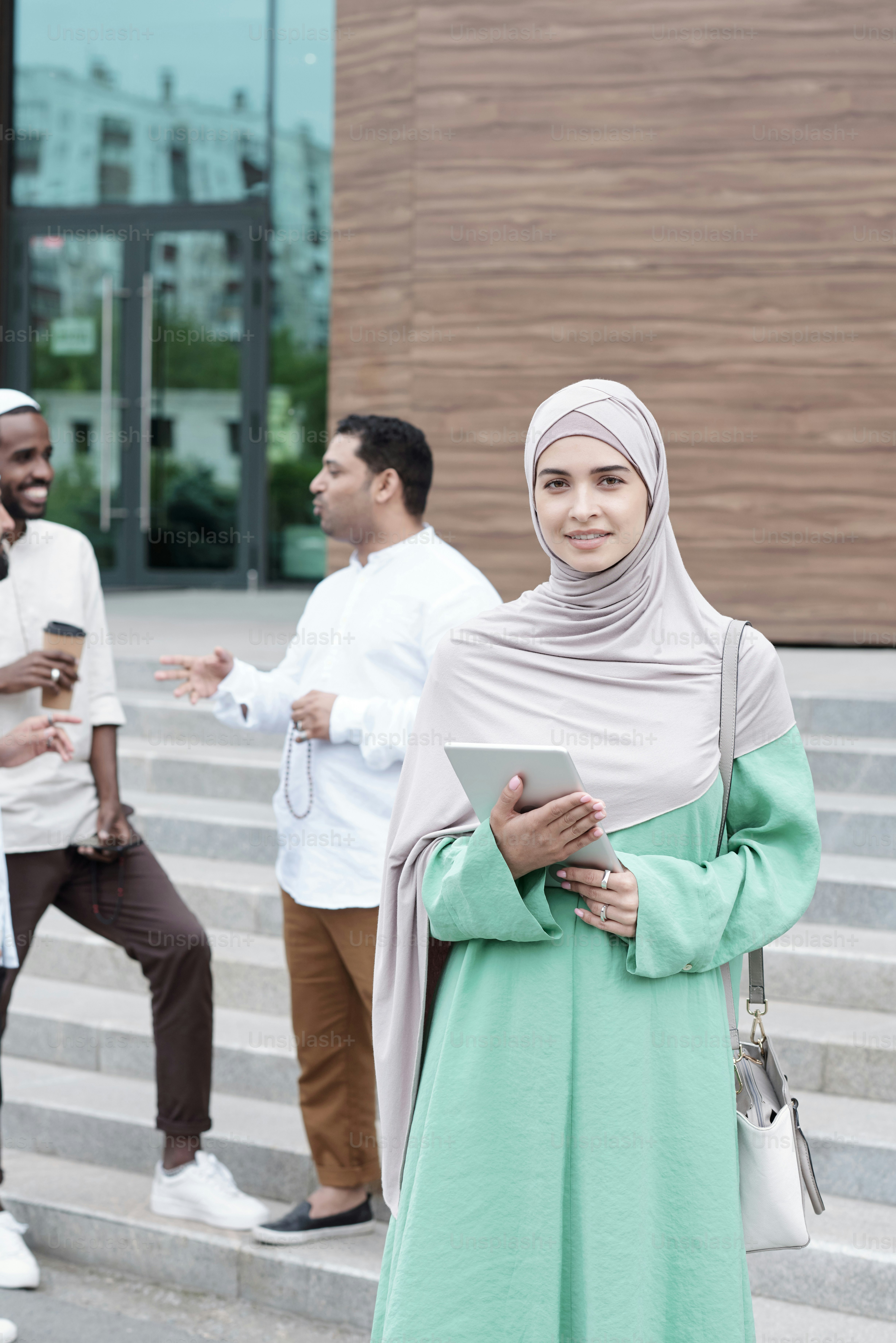 Portrait of modern young Islamic woman in hijab and national dress standing with tablet outdoors