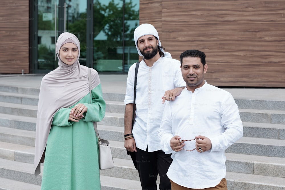 Grupo de jovens colegas muçulmanos de pé juntos contra o edifício de escritórios moderno e olhando para a câmera