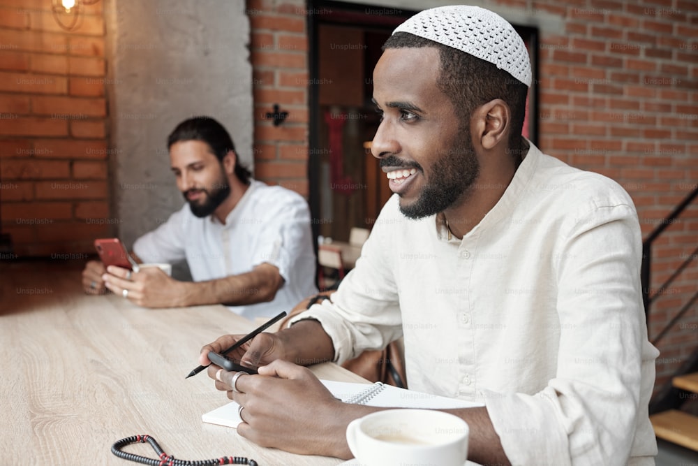 Joven negro sonriente con gorra musulmana sentado a la mesa y escribiendo ideas en el bloc de notas mientras envía mensajes de texto por teléfono