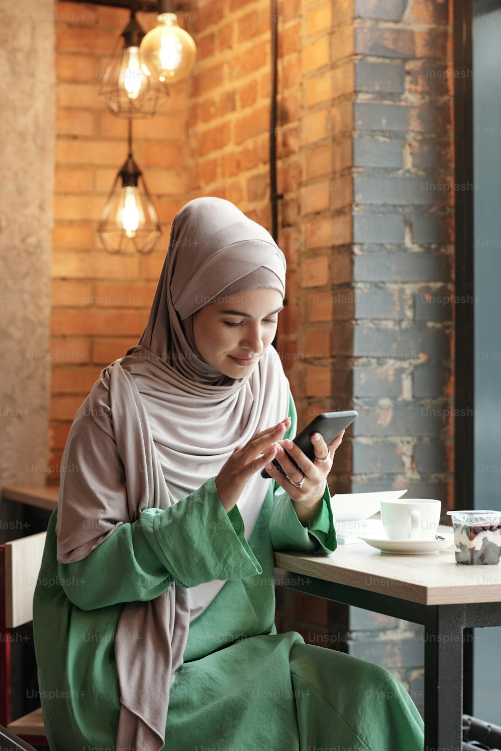 Contenu jeune femme musulmane en hijab assise à table et buvant du café tout en se connectant au WIFI dans un café