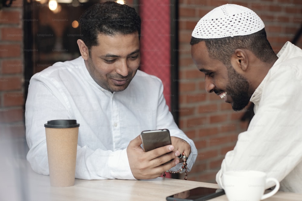 Positive young Muslim friends in white shirts sitting in cafe and watching photos on social media