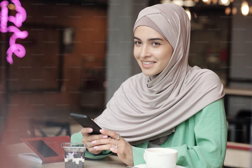 Jeune femme souriante et attirante en hijab assise à table dans un café et utilisant un gadget dans un café