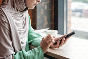 Close-up of calm young woman in hijab sitting at table and answering phone messages In cafe