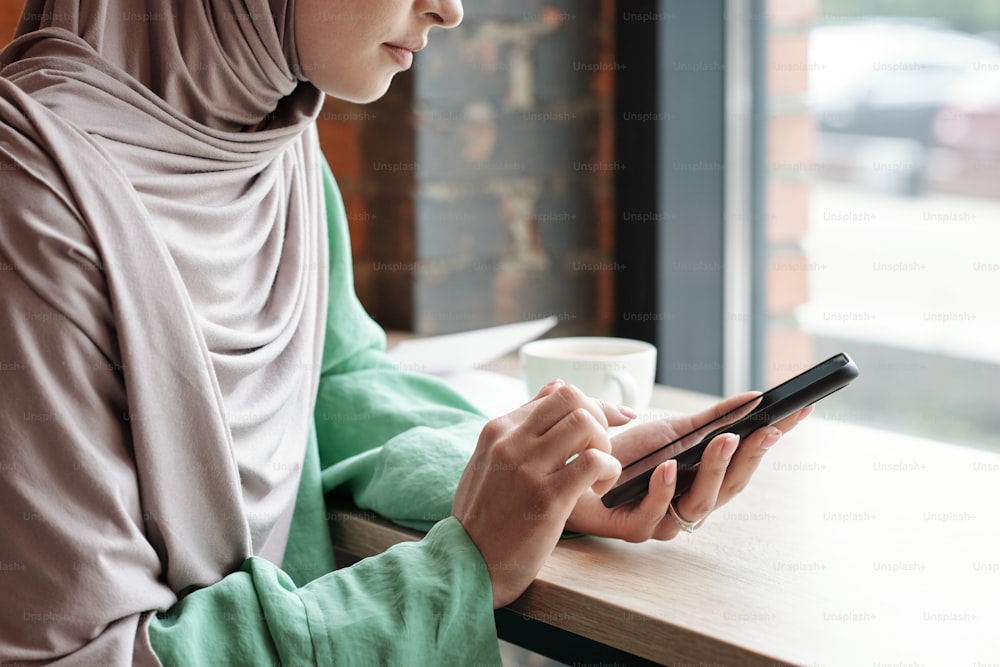 히잡을 쓴 차분한 젊은 여성이 테이블에 앉아 카페에서 전화 메시지에 응답하는 모습