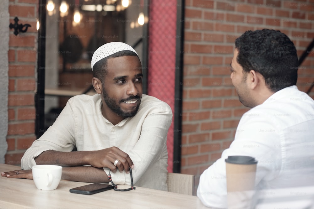 Smiling Black bearded man in kufi sitting with rosary beads at table and listening to Arabian friend in cafe