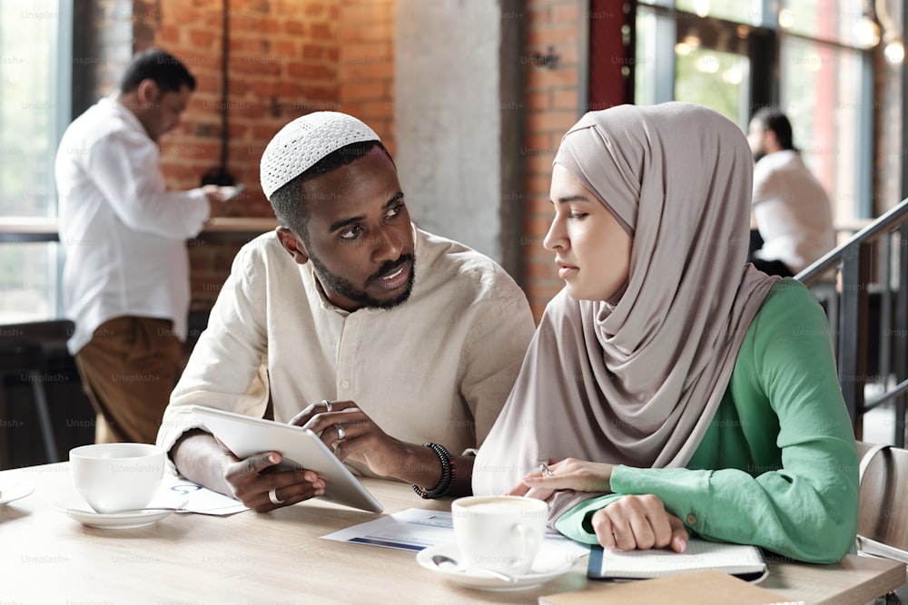 Young Black man in kufi cap using tablet while explaining data to female colleague in coffee shop