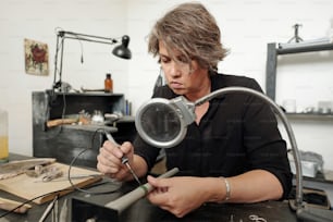 Concentrated female jeweler sitting in workshop with black furniture and soldering pieces of jewelry together under magnifying glass