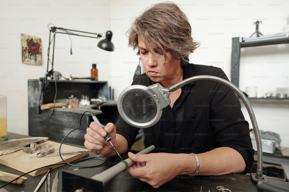 Concentrated female jeweler sitting in workshop with black furniture and soldering pieces of jewelry together under magnifying glass