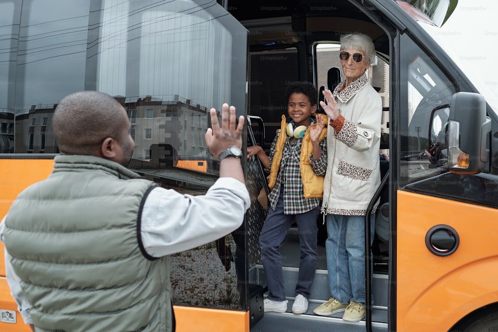 Menino negro sorridente abraçado pela avó acenando com a mão enquanto se despedia do pai, eles entram no ônibus