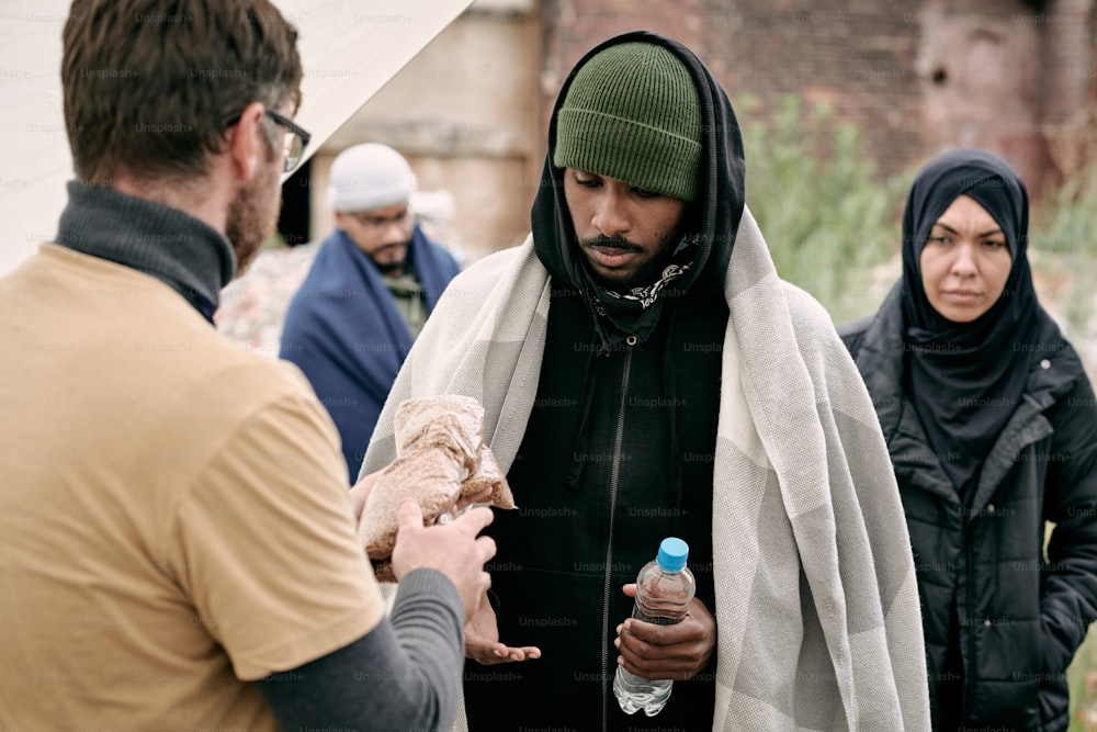 Social worker giving water and cereal to black man under plaid while providing food to refugees outdoors