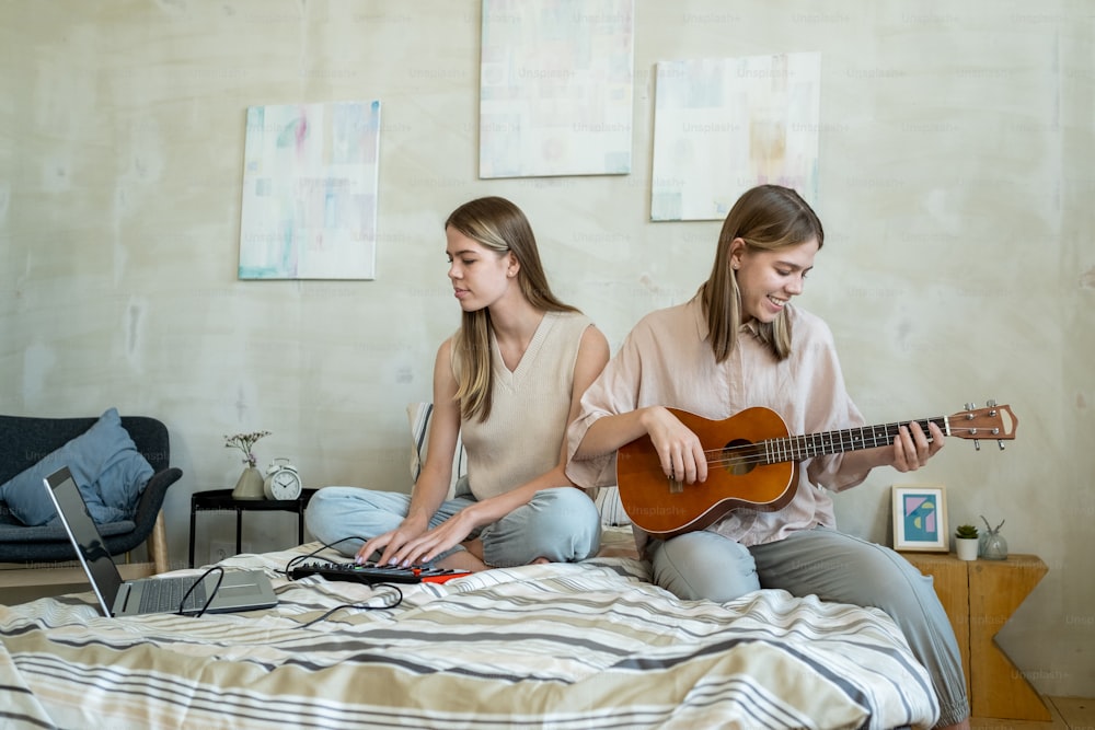 Cute teenage girl recording music of her twin sister playing guitar while both sitting on bed