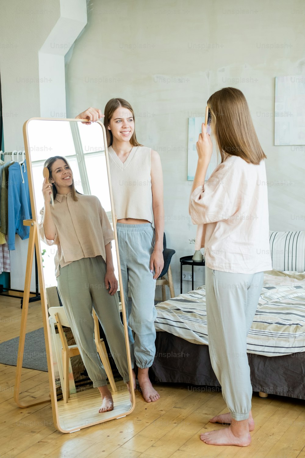 One of two cute twins brushing hair in front of mirror in bedroom while her sister looking at her