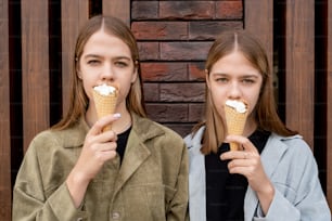 예쁜 쌍둥이가 건물 벽 앞에 서서 아이스크림 콘을 얼굴로 들고 있다