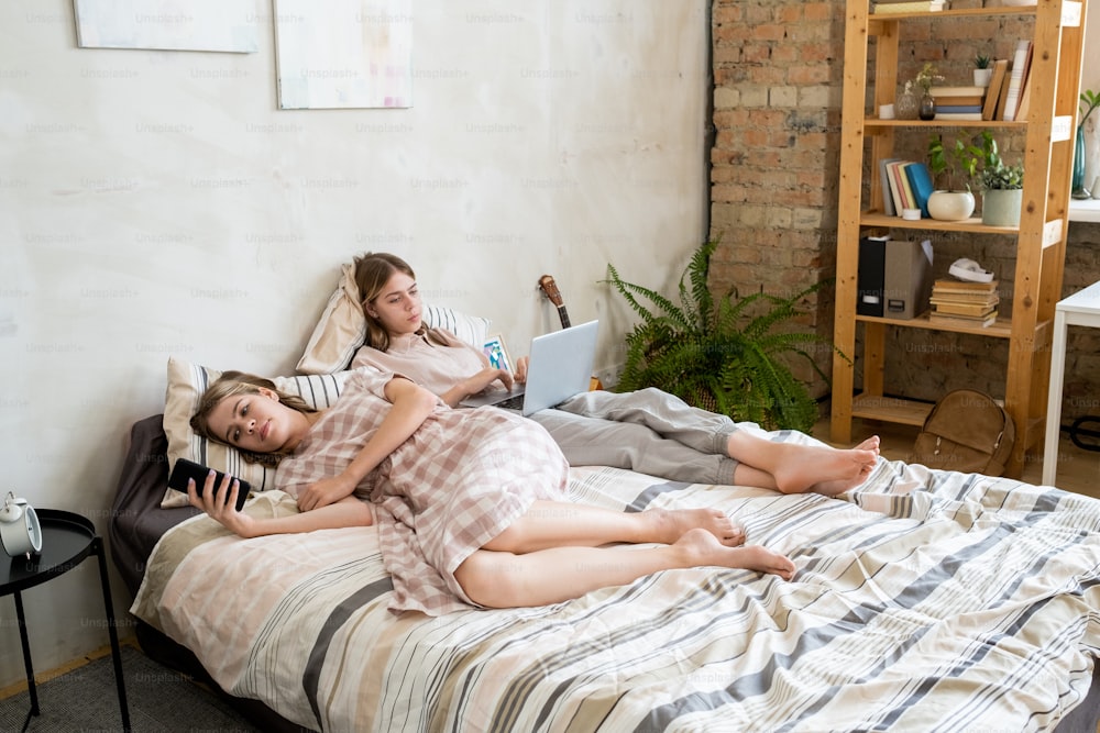 Two restful twin girls using mobile gadgets while lying on bed at lesiure
