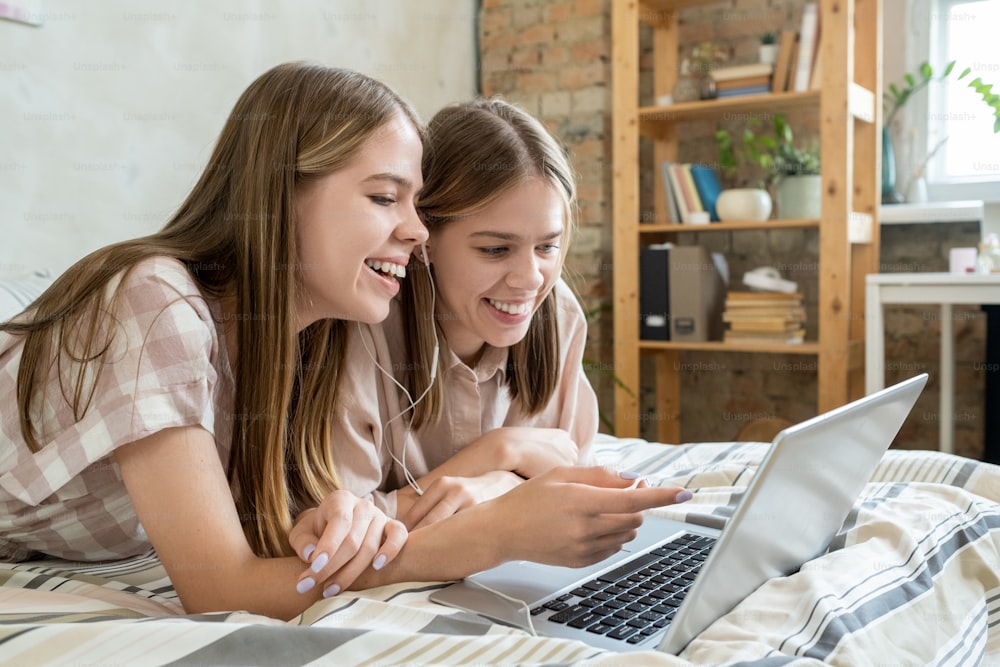 Two cheerful teenage girls discussing moment of online movie while relaxing on bed in front of laptop