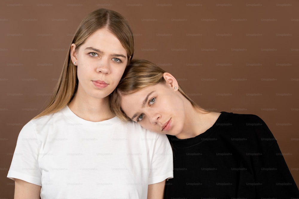 One of blond twin girls in white and black t-shirts keeping head on shoulder of her sister against brown background
