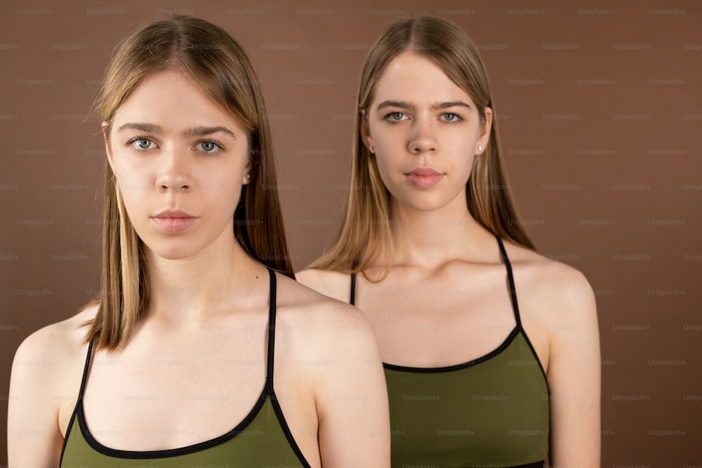 Gelassene Zwillinge in pistazienfarbener Unterwäsche, Tanktops, die in die Kamera schauen, während sie isoliert vor braunem Hintergrund stehen
