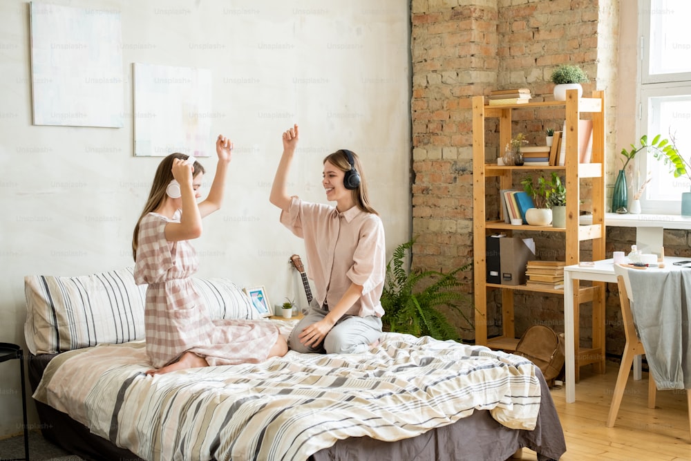 Excited teenage twin girls in headphones enjoying music and dancing at leisure while sitting on bed
