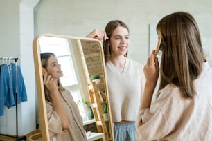 Happy teenage twins standing by large mirror and chatting while one of them brushing her long blond hair