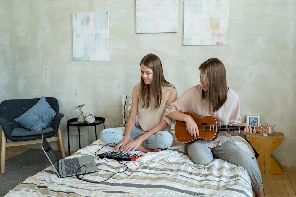 두 명의 행복한 십대 소녀가 집에서 음악을 녹음하는 동안 그 중 한 명은 기타를 연주하고 다른 한 명은 피아노 건반을 누르고 있습니다