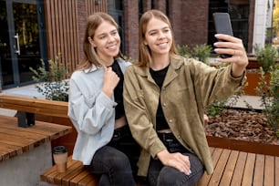 Happy adolescent twin girls looking at smartphone screen while resting on bench and making selfie in the street