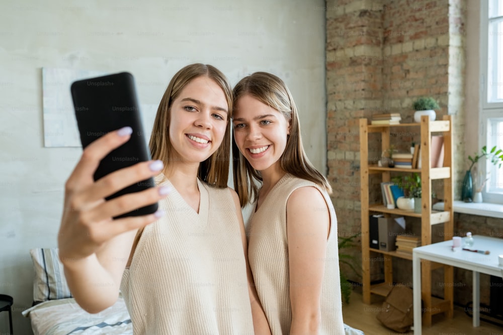 Happy twin girl with smartphone making selfie with her twin sister while both standing in front of camera in their bedroom