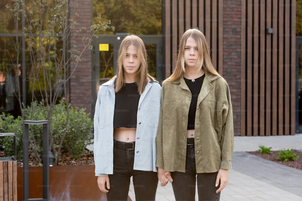 Ernsthafte weibliche Zwillinge im Teenageralter in Freizeitkleidung stehen vor der Kamera vor dem Gebäude
