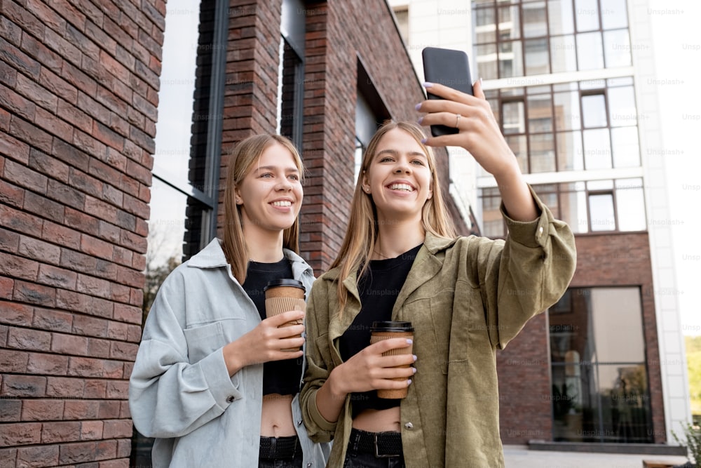 Smiling twin girls with drinks looking at smartphone camera while making selfie or communicating in video chat