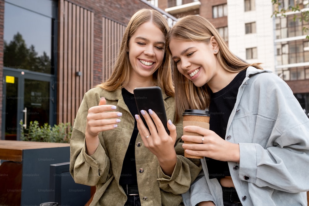 Junge Zwillingsschwestern in Freizeitkleidung schauen auf den Smartphone-Bildschirm, während sie sich entspannen und in der städtischen Umgebung etwas trinken