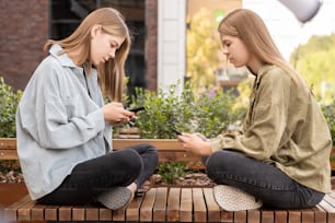 쌍둥이 소녀들은 서로 앞 벤치에 앉아 도시 환경에서 여름날 스마트폰을 스크롤한다